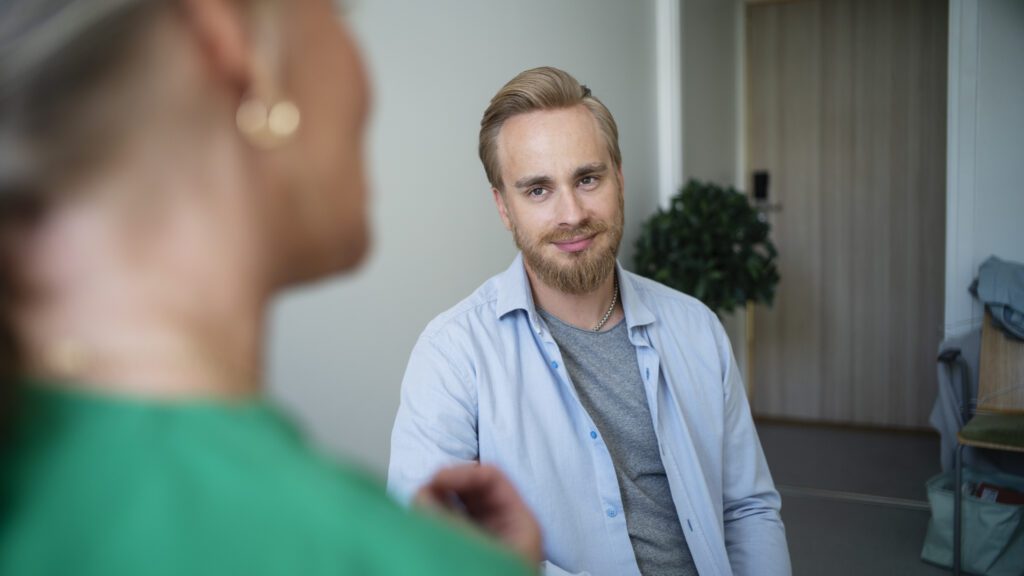 Vaaleahiuksinen mies, yllään vaaleansininen paita, hymyilee ystävällisesti katsoessaan keskustelukumppaniaan.